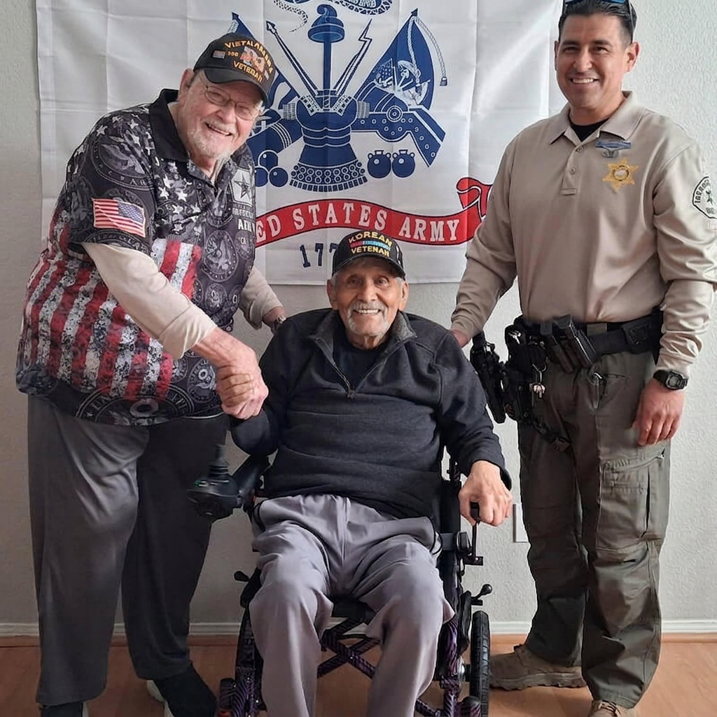 Robert Paiz smiling in a new electric wheelchair. He is accompanied by John Meyer, president of AARV, to the left wearing Veteran hat and an American flag shirt. A uniformed officer is on his other side. All three are standing in front of a U.S. Army backdrop.